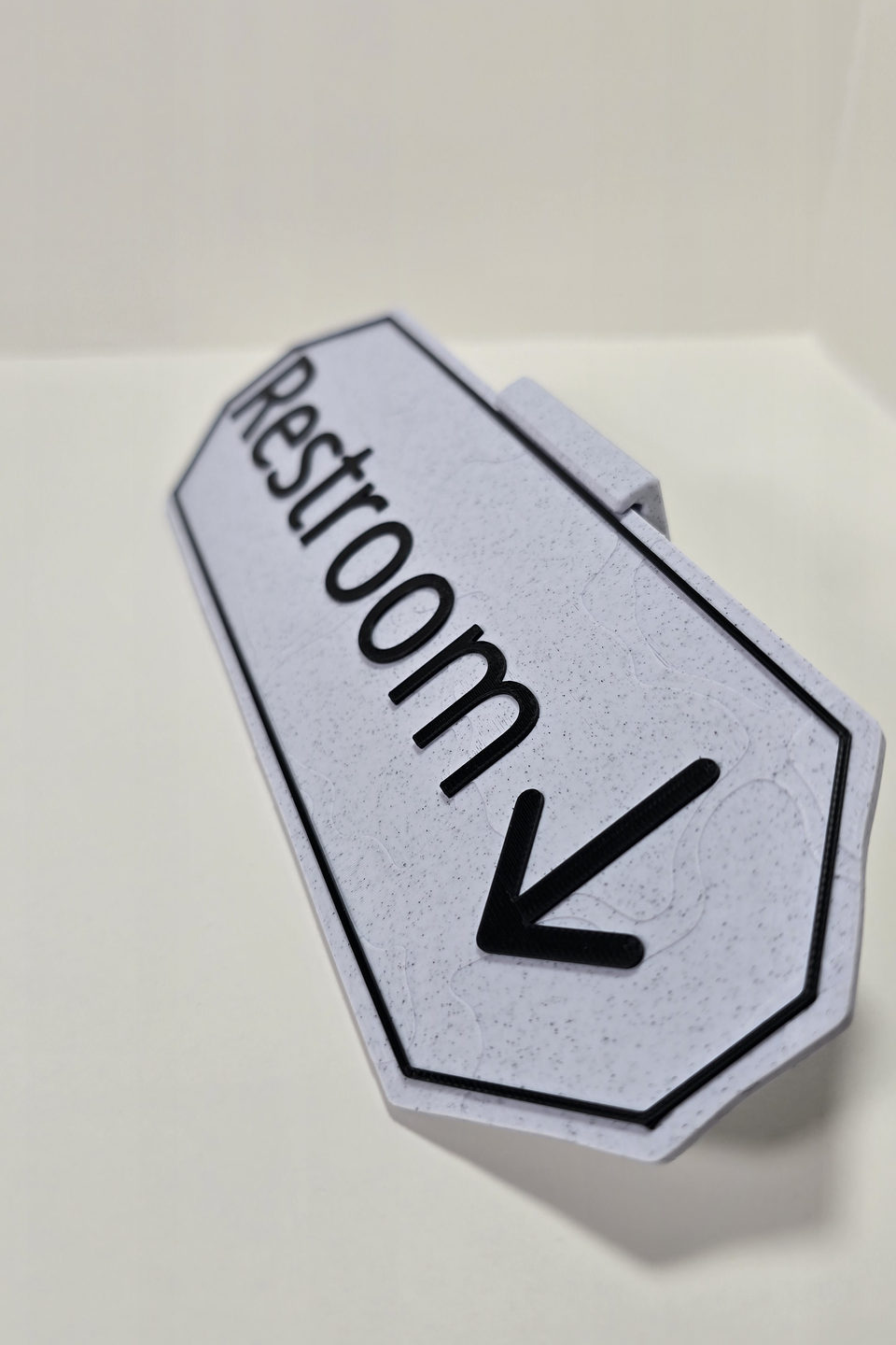 Close view looking down a Marble White Restroom sign showing its black text, arrow, border, angled corners, surface, and stand fit.