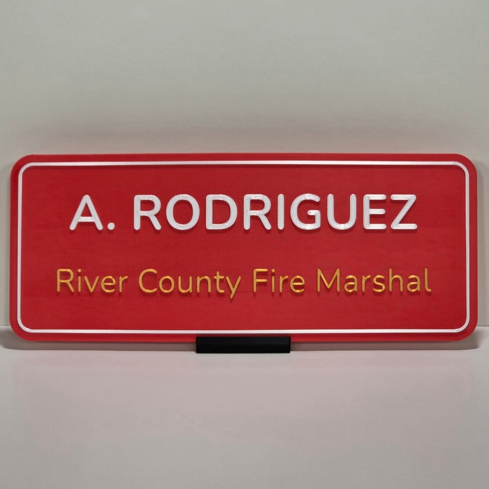 Red two-line office nameplate reading A. Rodriguez and River County Fire Marshal on a black tabletop stand.