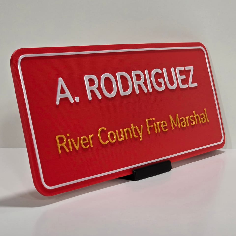Angled red office nameplate reading A. Rodriguez and River County Fire Marshal on a black tabletop stand.