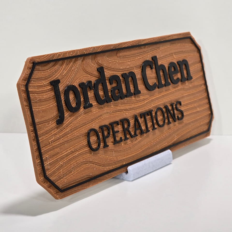 Angled wood-look office nameplate reading Jordan Chen and Operations on a marble-white tabletop stand.