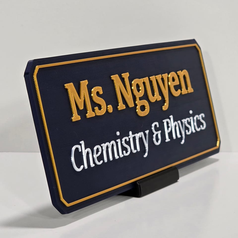 Angled navy teacher nameplate reading Ms. Nguyen and Chemistry & Physics on a black tabletop stand.